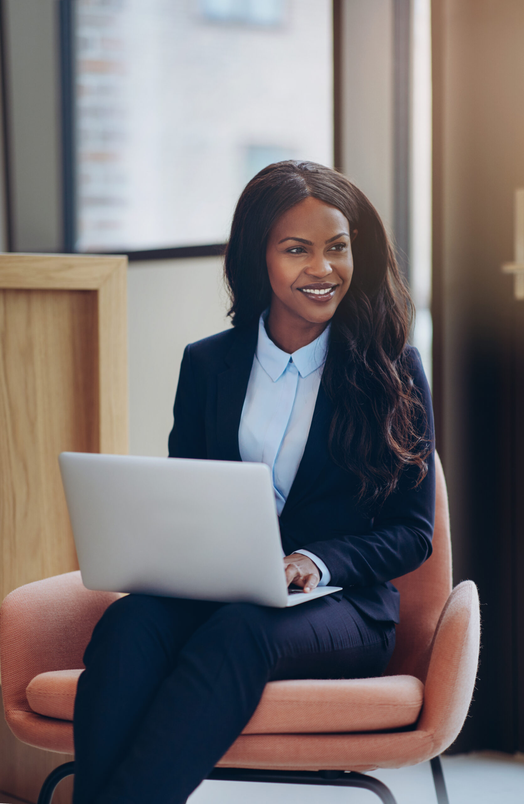 Smiling young African American businesswoman sitting in a chair in an office lounge working online with a laptop